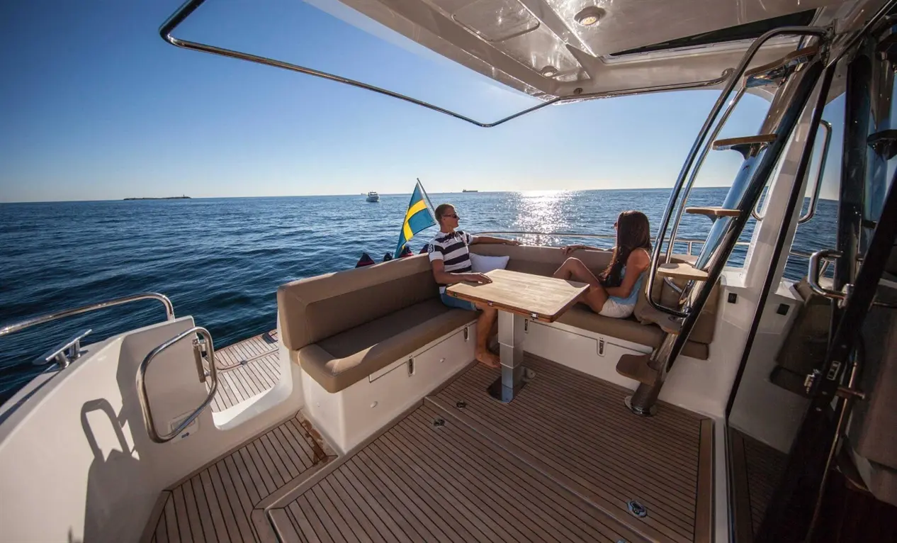 A couple relaxes on a yacht deck by the sea, with a table between them and a Swedish flag visible in the background.